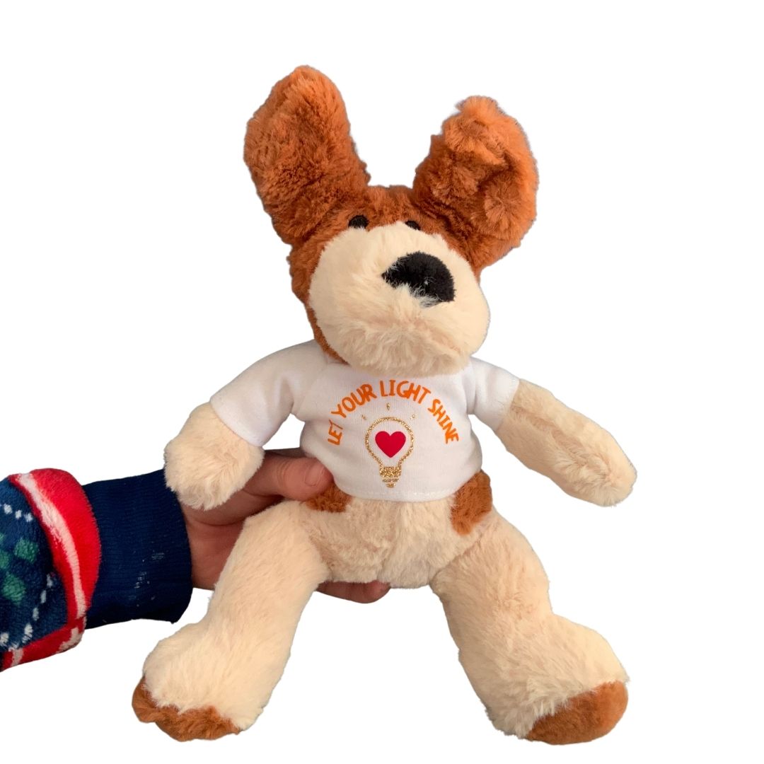 A photograph of a child’s arm and hand holding a totally ‘pawsome’ Mary, Martha & Meg soft toy dog. This small plush pup is cream with light brown ears and features. He is wearing a white t-shirt with the words, 'Let your light shine.' This positive encouragement can be given to any child, including those who are still young at heart. The orange text is curved above a design of a gold glitter lightbulb with a red heart at its centre. 