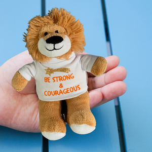 This adorable Mary, Martha & Meg soft toy is a small, soft lion called Louis. He is light brown and is wearing a white t-shirt with the encouraging words ‘Be strong and courageous,’ in orange, ironed-on text. There is a gold glitter shooting star above this verse from the Bible (Joshua 1:9).  Louis is held in an open hand. The background of the photograph shows blue wooden boards. 