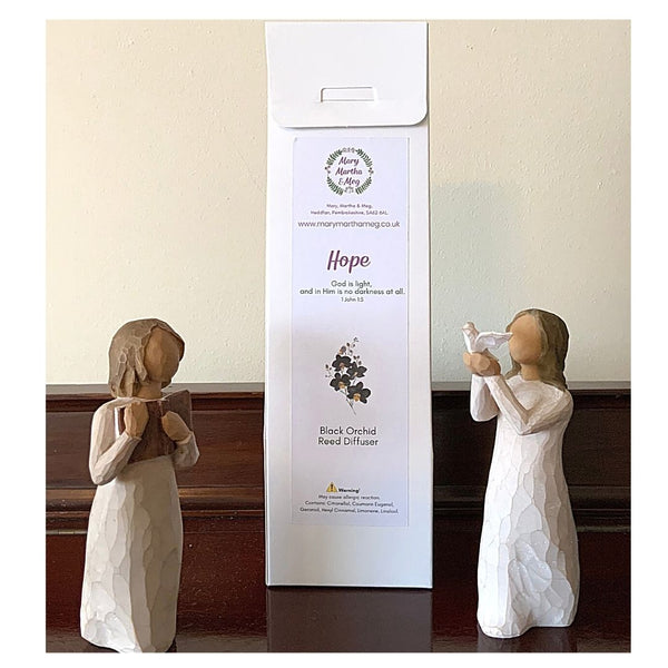 A photograph showing a Mary, Martha and Meg ‘Hope’ Reed Diffuser in its white packaging. The back of the white packaging box has the Mary, Martha & Meg logo. Written text includes the name of the diffuser ‘Hope,’ the Bible verse and reference which inspired this fragrance, the name of the fragrance, ‘Black Orchid’ as well as an allergy warning.  There is an illustration of black orchids. The reed diffuser packaging is placed between two wooden figurines, for illustration purposes only.