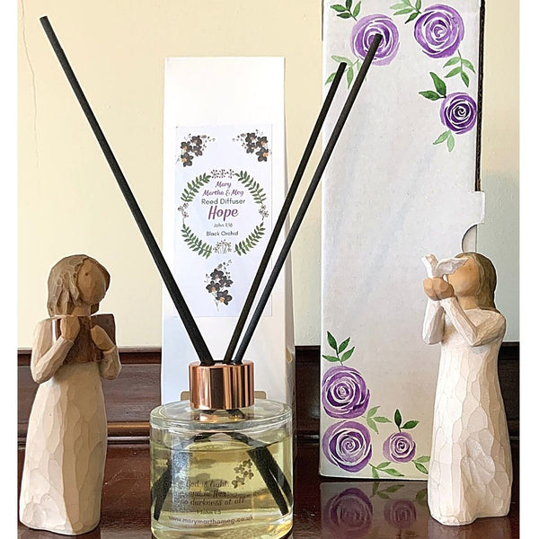 A photograph showing a Mary, Martha and Meg Hope reed diffuser bottle, a white packaging box for the diffuser and a hand-painted white box of purple roses. Two wooden figures are seen either side of the Mary, Martha & Meg products, for illustration purposes only. One is holding a book, the other a dove.