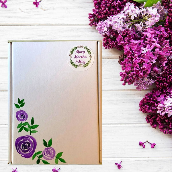 A photograph of a hand-painted Mary, Martha & Meg mini gift letter box. Using acrylic paints, Meg has painted purple roses. A Mary, Martha & Meg logo sticker is placed in the top right corner. The pretty giftbox is pictured on a white wooden table with lilac flowers in the background.