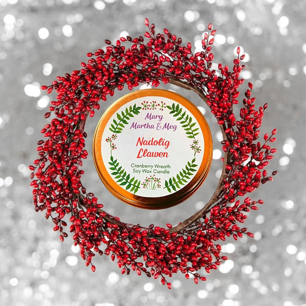 A Mary, Martha & Meg rose gold candle lid. The white label has the name of the candle, Nadolig Llawen, in red text in the centre of the label. Underneath, in green text, is the name of the fragrance, Cranberry Wreath, as well as the text ‘Soy Wax Candle.’ All text is framed by the Mary, Martha and Meg logo of a green wreath of leaves with tiny flowers. The candle lid sits inside a wreath of red cranberries. Behind this is a silver background. 