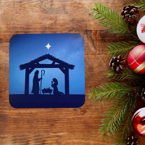 A pretty Christmas coaster created by Mary, Martha & Meg. The square coaster has rounded edges. The design is of a Nativity scene showing the silhouettes of Mary and Joseph in a stable, worshipping baby Jesus lying in a manger. The scene is captured against an inky blue sky with a white star shining above the stable. The coaster is photographed on rich brown wood with pine cones, leaves and red and white Christmas decorations to the right-hand side of the photograph.