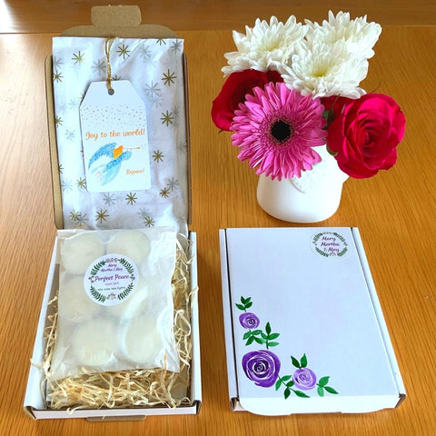 A photograph showing a filled mini letter gift box, a closed painted gift box and a vase of flowers. The mini letter box has Christmas tissue paper and natural shredded filler and a packet of 6 peace tealights with a logo sticker. A gift tag of an angel hangs on the inside cover. The closed gift box shows purple roses hand-painted by Meg, with a small Mary, Martha & Meg logo sticker in the top right corner. A lovely gift which can be posted straight to you or to a friend, and which fits through a letterbox.