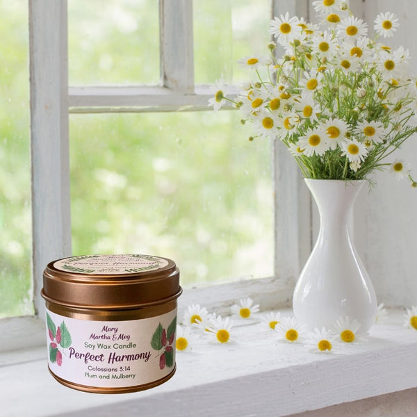 A Mary, Martha & Meg soy wax scented small candle tin pictured on a white windowsill next to a vase of daisies, with individual daisies scattered next to the candle. The candle name, Perfect Harmony, is accompanied by the Bible reference Colossians 3:14. The natural soy wax is fragranced with the beautiful floral notes of plum and mulberry.