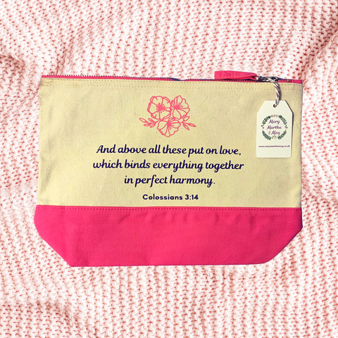 A photograph of a natural canvas zip pouch with a pink zip and pink base. The Bible verse reads, "And above all these put on love, which binds everything together in perfect harmony." Colossians 3:14. Above the text is a trio of pink flowers and petite leaves. A Mary, Martha & Meg tag can be seen hanging from the zipper on the right of the pouch. This beautiful item is pictured on a pale pink crocheted blanket.