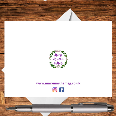 A photograph showing the reverse of a Just to Say greetings card. In the centre is the circular Mary, Martha & Meg logo of green leaves and tiny purple flowerheads, surrounding the Mary, Martha & Meg purple text. Beneath this, also in purple text, is a reference to this small business website: www.marymarthameg.co.uk The Pinterest logo and Facebook logo is also included. The edge of a white envelope can be seen behind the Just to Say card, which is placed on a wooden writing surface with a silver pen.
