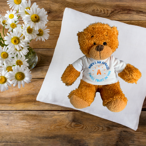 This delightful Mary, Martha & Meg Prayer Teddy Bear is pictured resting on a white pillow against a dark brown wooden background, with large white daisies to the left of the photograph. The adorable, soft fur prayer teddy bear has a friendly face and wears a t-shirt with the beautiful verse from Psalm 56:3, "When I am afraid, I will trust in you." The carefully hand-printed text is written in two shades of blue in a circle, with orange praying hands in the centre.