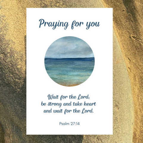 A Mary, Martha & Meg encouragement card with the words, Praying for You,’ above a circular painting of the sea, which has been printed from an original watercolour painting by Meg. The encouraging Bible verse underneath the art is, “Wait for the Lord, be strong and take heart and wait for the Lord,’ Psalm 27:14. The card is photographed on a sandy beach near Meg’s home.