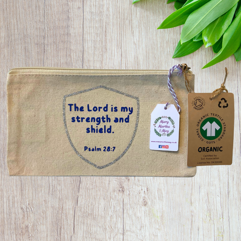A Mary, Martha & Meg organic, natural, canvas zip pouch featuring the encouraging Bible verse, ‘The Lord is my strength and shield, Psalm 28:7.’ The Bible verse is printed in blue, inside a silver glitter shield. The design has been hand-printed by Meg. A Mary, Martha & Meg tag hangs from the zip, as well as a label showing the organic credentials of the zip pouch. The zip pouch is pictured on a wooden table with green leaves in the top right corner. 