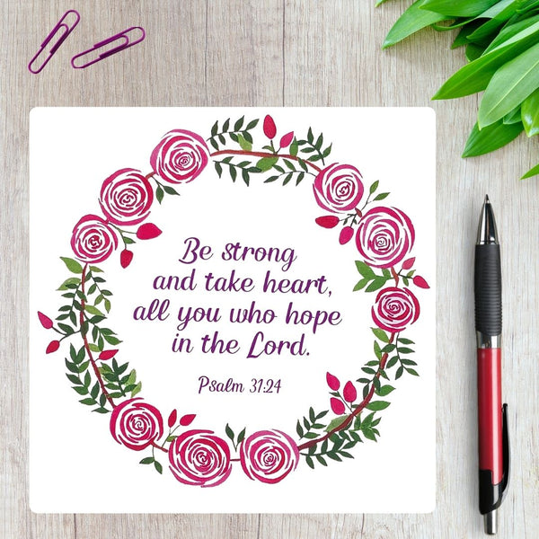 A square greeting card with the encouraging words from Psalm 31:24  surrounded with a pink rose wreath and foliage. The card is on a light wooden background  with red paperclips, next to a red pen. and green leaves in the corner.