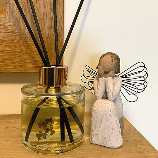 A Mary, Martha & Meg luxury reed diffuser bottle with a rose gold coloured top is seen placed on a wooden mantlepiece below a wooden-framed mirror. The bottle design shows a spray of black orchids, referencing the fragrance of this luxury diffuser scent. To the right of the diffuser is a wooden sitting angel, which is used for illustration purposes only. 