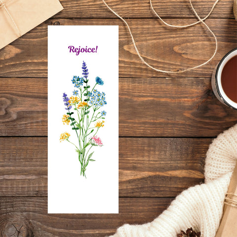 A Mary Martha & Meg white bookmark featuring the word ‘Rejoice’ above a floral bouquet of pretty wildflowers. A perfect size to pop into any book, notebook, Bible or handbag. The bookmark is photographed on a brown wooden table. In the top left corner the edge of a brown parcel can be seen. In the top right corner there is some loose string. A cup of coffee and a cream jumper can be seen on the right side of the photo.