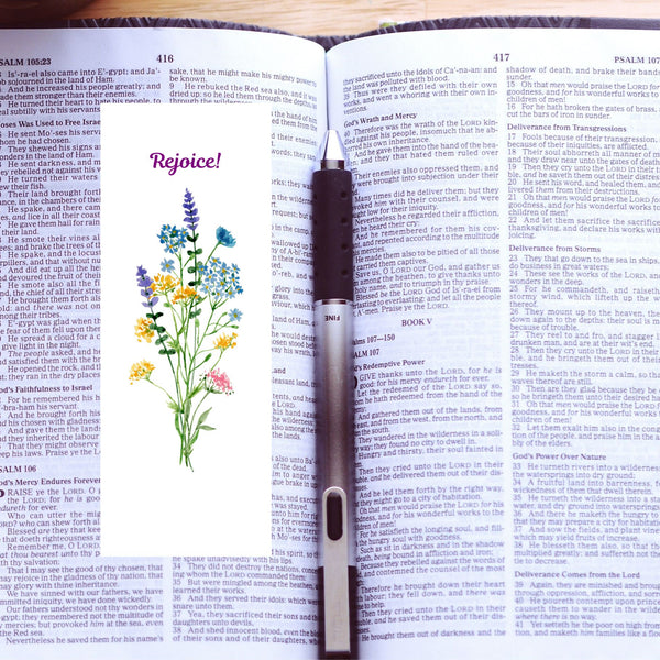 A Mary Martha & Meg white bookmark featuring the word ‘Rejoice’ above a floral bouquet of pretty wildflowers. A perfect size to pop into any book, notebook, Bible or handbag. The bookmark is photographed on an open Bible with a black and silver pen to the right of the bookmark.
