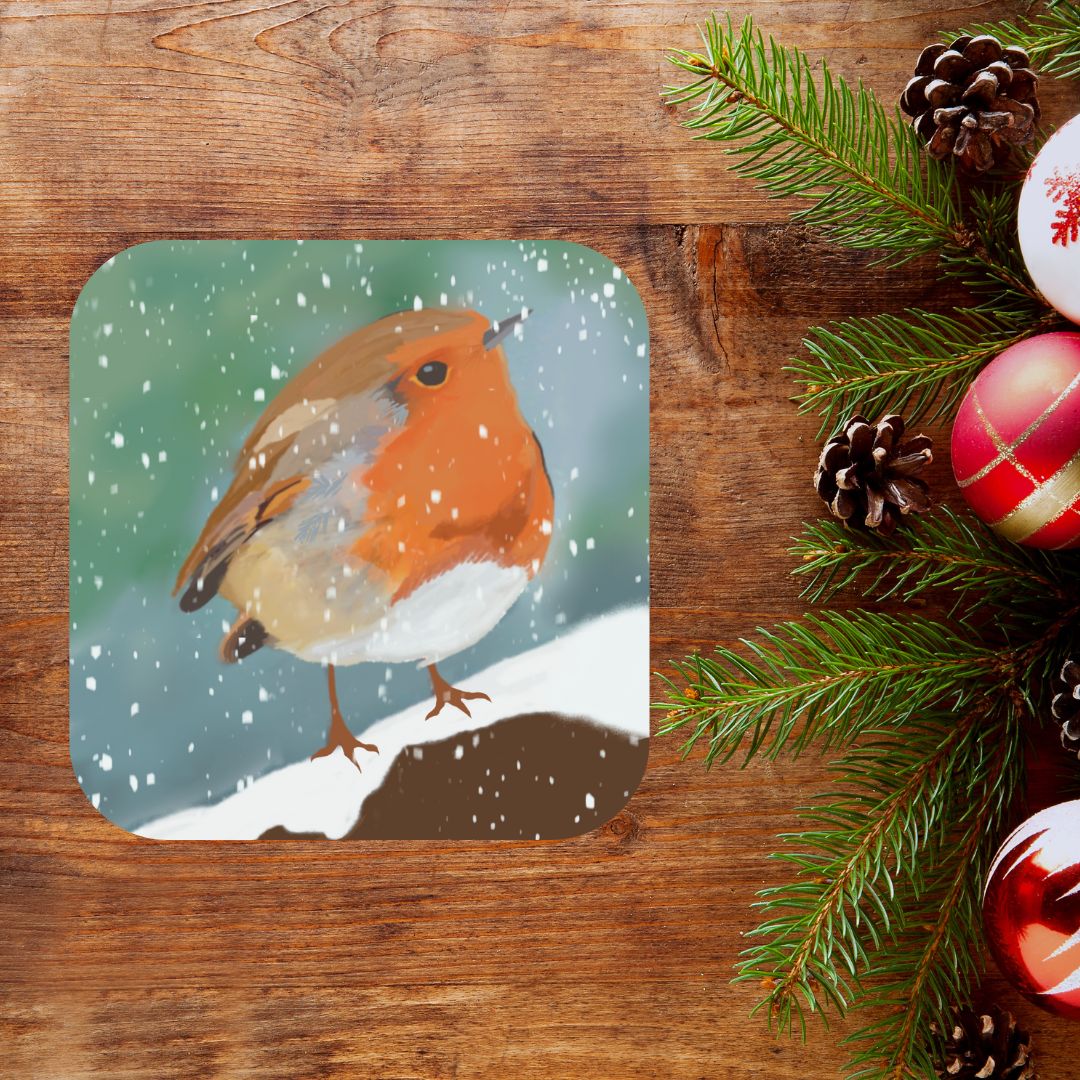 A photograph of a Mary, Martha & Meg pretty Christmas coaster. The design centres on a traditional winter scene of a robin perched on a snowy branch. The coaster is pictured against a rich wooden background with festive decorations on the right-hand side.