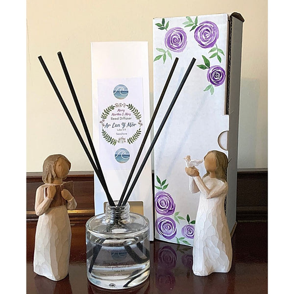 A photograph showing a Mary, Martha and Meg Seashore reed diffuser bottle, a white packaging box for the diffuser and a hand-painted white box of purple roses. Two wooden figures are seen either side of the Mary, Martha & Meg products, for illustration purposes only. One is holding a book, the other a dove.