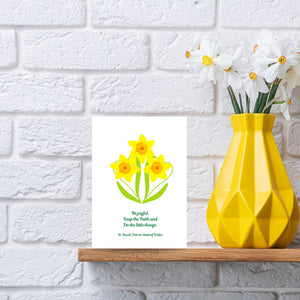 A bright, eye-catching card of three golden daffodils features the last words of David, Patron Saint of Wales, “Be Joyful. Keep the Faith. Do the little things.” It is a lovely reminder to serve each other in the little things we do, no matter how small. This Mary, Martha & Meg card is pictured next to a yellow vase of white daffodils, which sit on a wooden shelf against a white brick wall.