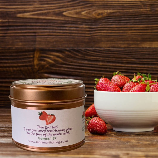 Showing the back of Mary, Martha & Meg Strawberry Delight Candle. Text from Genesis 1:29, '"Then God said, "I give you every seed-bearing plant on the face of the whole earth."' A white bowl of strawberries are behind the candle. The background of the picture is dark varnished wood.