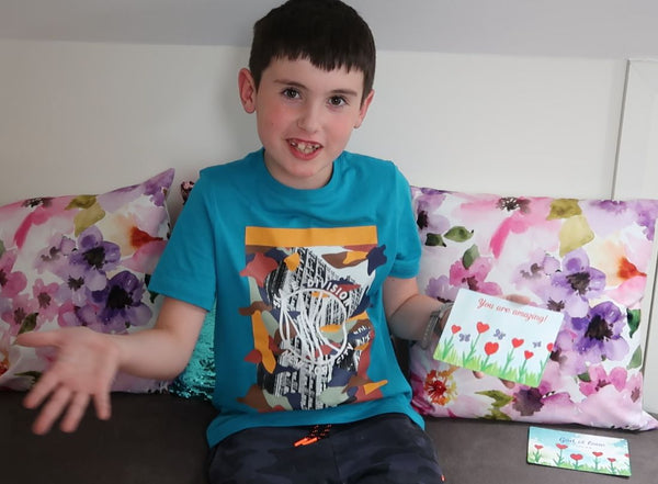 A short video of a happy child talking about the postcard he has created. He is showing his hand-painted watercolour postcard in his hand. Pastel floral cushions can be seen behind him. He is sitting on a grey sofa. 