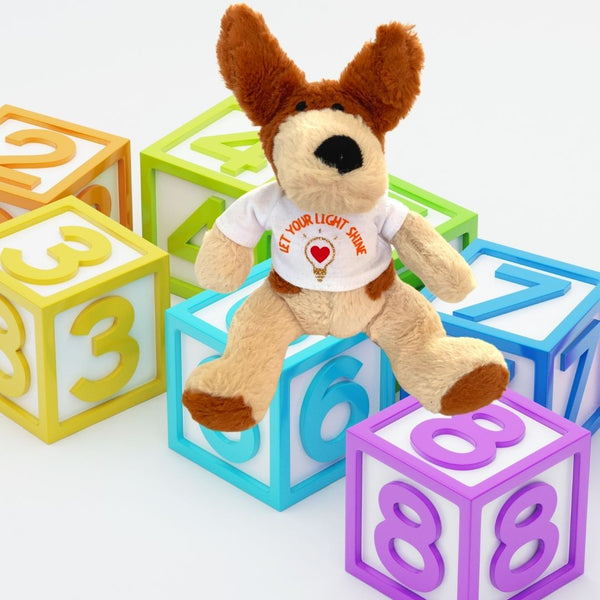 An adorable Mary, Martha & Meg soft toy dog sitting on brightly coloured nursery play cubes with numbers on them This small plush pup is cream with light brown ears and features. He is wearing a white t-shirt with the words, 'Let your light shine.' This positive encouragement can be given to any child, including those who are still young at heart. The orange text is curved above a design of a gold glitter lightbulb with a red heart at its centre. 