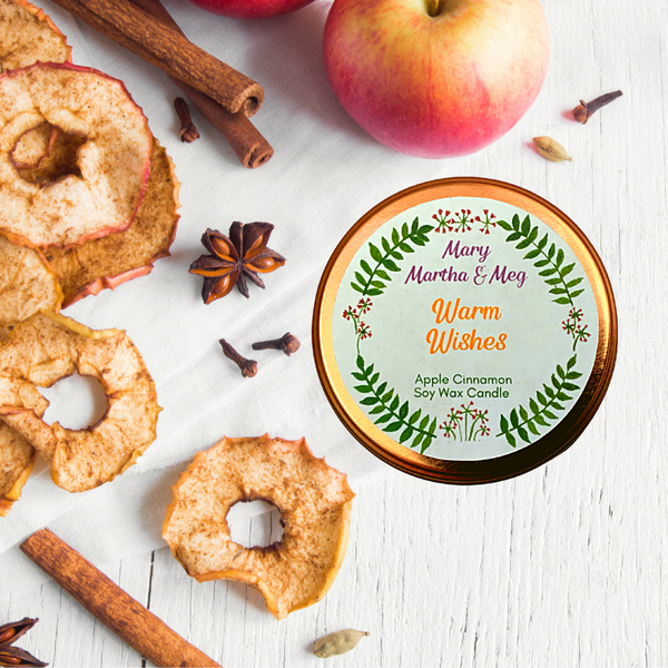 An aerial view of a Mary, Martha & Meg  'Warm Wishes' Apple Cinnamon Soy wax candle in a rose gold tin.  The candle is surrounded by dried apple slices, whole red apple and cinnamon sticks.