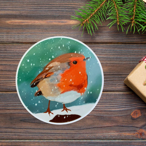 A photograph showing a Mary, Martha & Meg coaster of a winter robin. The ceramic white edge of the circular coaster can be seen. The robin is perched on a snow-covered branch. The greeny-blue sky has snowflakes falling. The coaster is photographed on dark wood. Green pine leaves and the tip of a Christmas gift can be seen to the right of this lovely winter robin coaster.