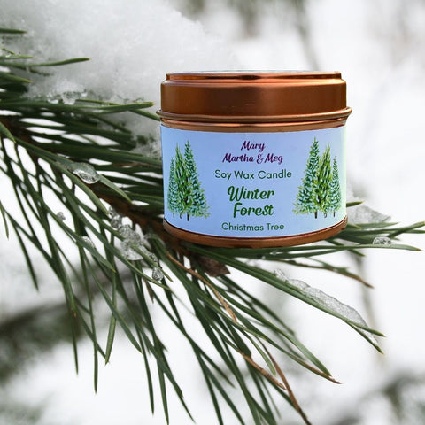 A photograph of a seasonal Winter soy wax candle created by Mary, Martha & Meg. The name of the candle, ‘Winter Forest’ is in green text. Underneath, also in green text, is the name of the fragrance, ‘Christmas Tree. The front of the Winter Forest soy wax candle label is decorated with fir trees. This beautifully scented soy candle is pictured on the branch of a forest fir tree.  