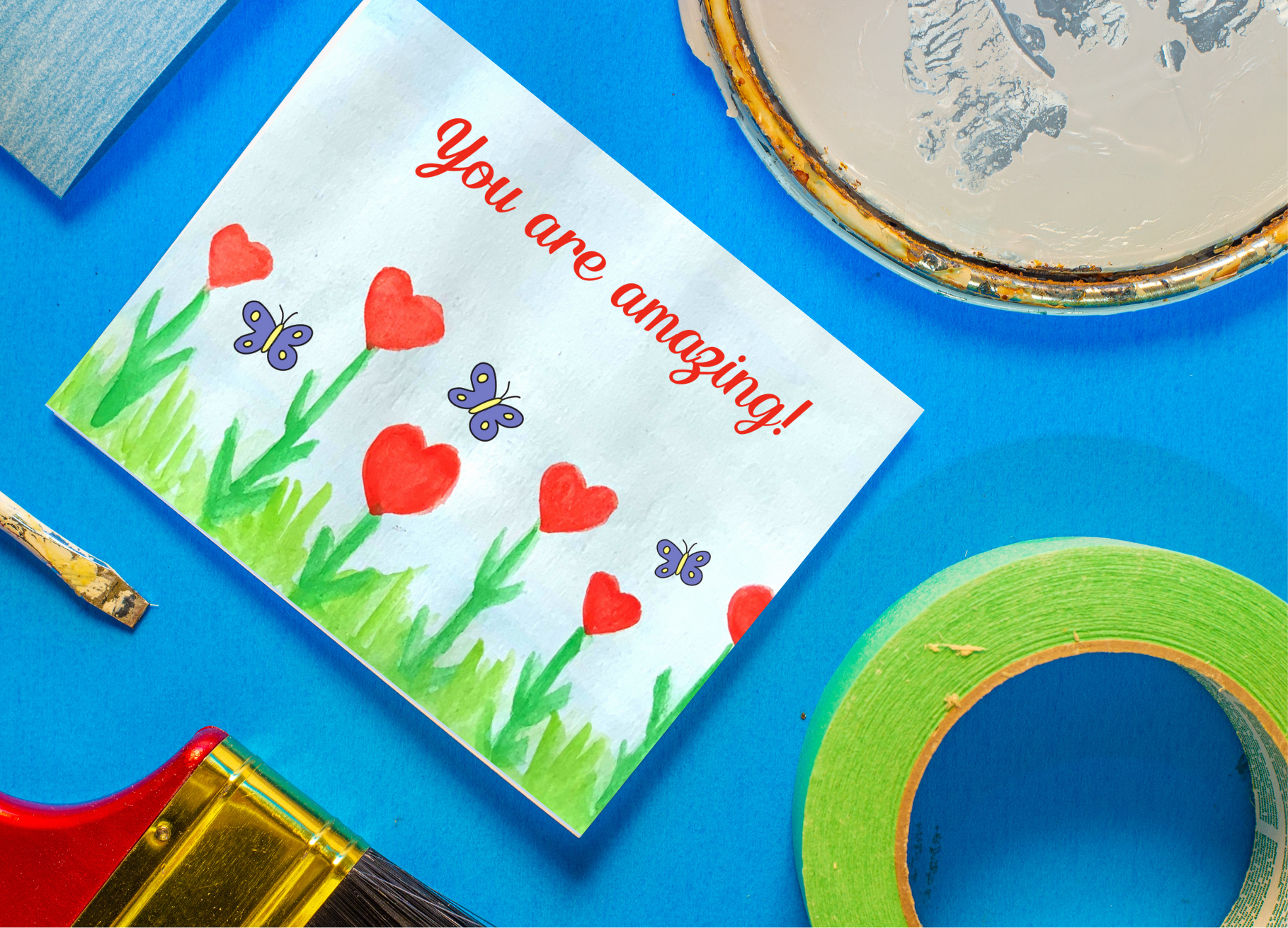 A photograph of a postcard with the words ‘You are amazing.’ The design is of red heart flowers growing between blades of green grass. Purple and yellow butterflies flutter in the blue sky. This watercolour has been hand-painted by a young child. The background of the photographs is blue. There are paint items surrounding the photo (card, a paint tub lid, green tape, a red handled brush and a smaller paintbrush).