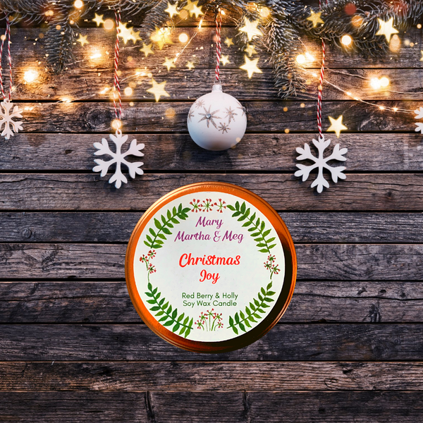 A photograph of the lid of a Mary, Martha & Meg soy wax candle. The name of the candle, 'Christmas Joy,' is written in a red font. The rose gold lid is pictured on dark wood with white Christmas decorations and warm lights above it